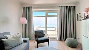 A modern living room with a sofa, armchair, and ottoman, a lamp, and a view of the sea through large windows.