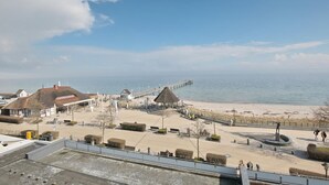 A coastal promenade with a pier extending into the sea, a beach, and buildings with thatched roofs.