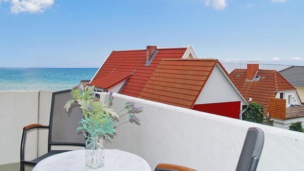 A balcony with a table and chairs, overlooking a coastal area with red-roofed buildings and the sea. - Strandperle Apartment 201 - Strandperle (Scharbeutz)