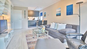 A modern living room with a grey sofa, a glass coffee table, and a flat-screen TV. There is a kitchen in the background with grey cabinets and a dining area. - Strandperle Apartment 201 - Strandperle (Scharbeutz)