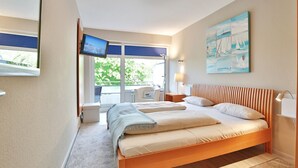 A bedroom with a large bed, a TV mounted on the wall, a window with a view of greenery, and a painting on the wall. - Strandperle Apartment 201 - Strandperle (Scharbeutz)