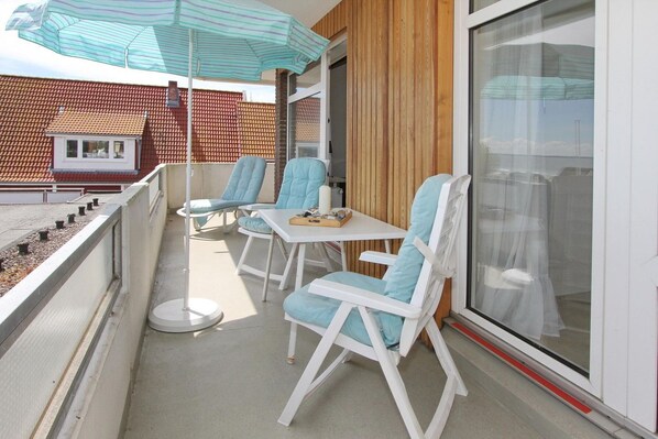 A balcony with white chairs and a table, a striped umbrella, and a view of a building with a tiled roof.