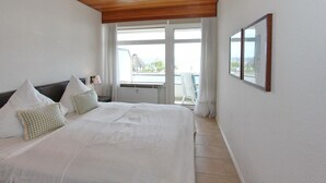 A bedroom with a large bed, two pillows, a nightstand, and a window with a view of a building and trees. - Strandperle Apartment 107 - Strandperle (Scharbeutz)