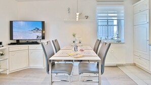 Un área de comedor moderna con una mesa grande, seis sillas, un televisor de pantalla plana mostrando una escena de playa y una ventana con persianas.