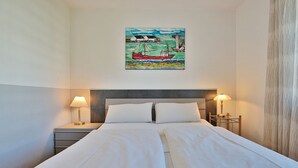 A double bed with white linens, two bedside tables with lamps, and a painting of a sailboat on the wall.