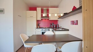 A compact kitchen with white cabinets, a dark countertop, and a red backsplash. There are two white chairs at the table, and a coffee machine is visible.
