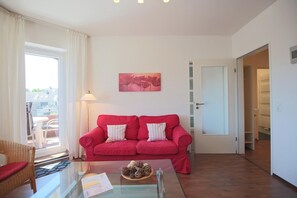 Sala de estar com um sofá vermelho, mesa de centro de vidro e uma cadeira de vime. Há uma porta que dá para um armário e uma vista para o exterior pela janela.