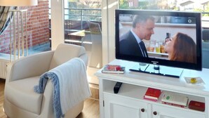 A modern living room with a flat-screen TV, a sofa, a lamp, and a view of a residential area through large windows.