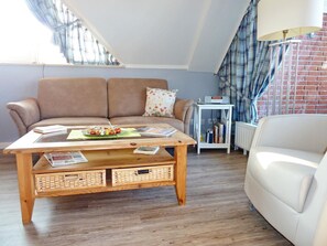 A cozy living room with a brown sofa, a wooden coffee table with magazines and a plate of food, a white armchair, and a small white side table with a radio.