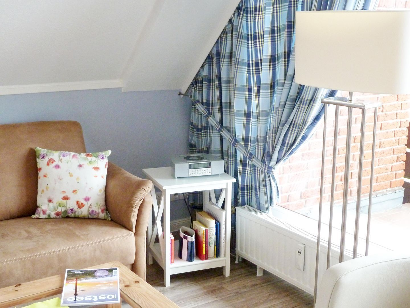 A cozy living room with a sofa, a small table with a magazine, a bookshelf, a radiator, and a window with plaid curtains.