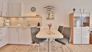 Une cuisine moderne avec des meubles blancs, une table à manger avec deux chaises, une machine à café et une horloge au mur.