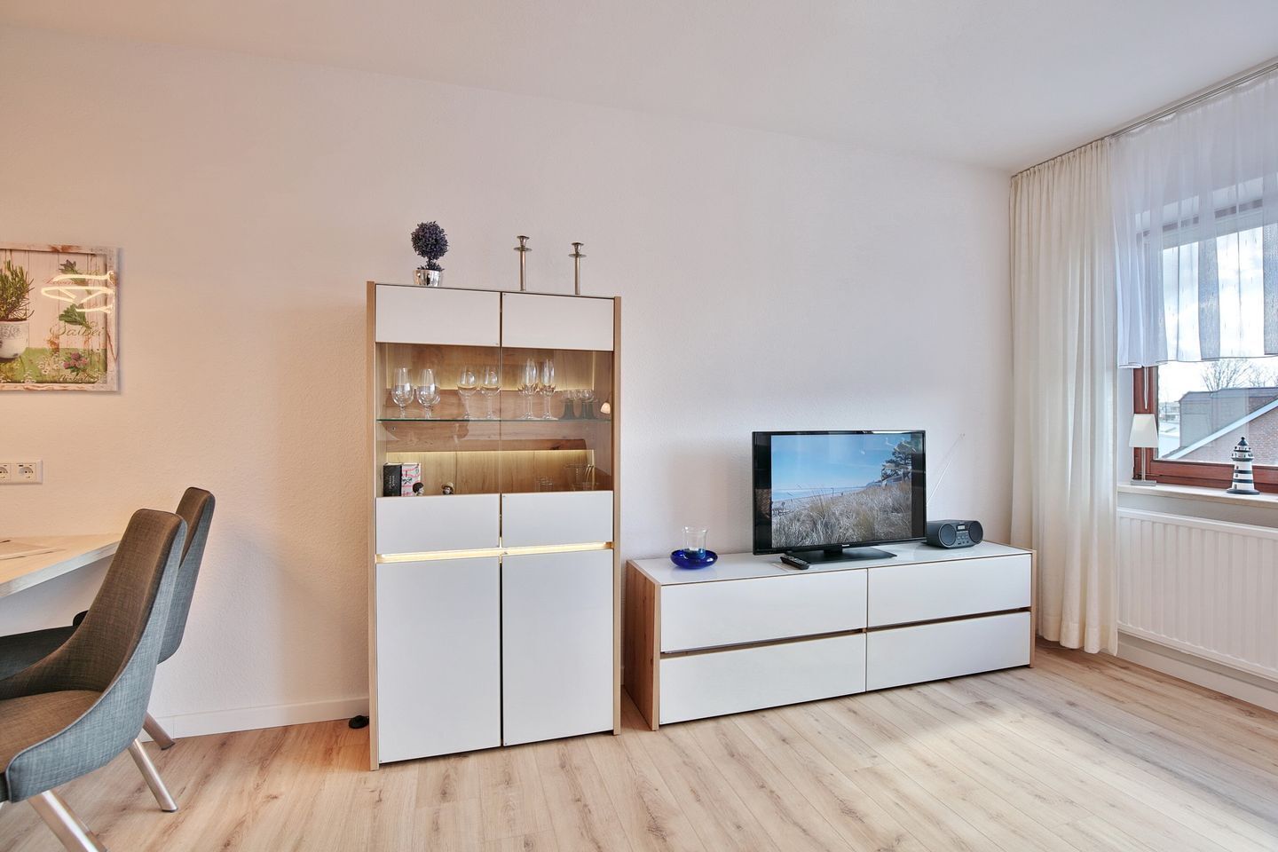 Un salon moderne doté d’un téléviseur à écran plat, d’un meuble vitré contenant des verres à vin et d’un coin repas avec une chaise.