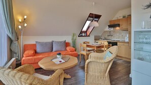 A modern living room with a sofa, coffee table, and wicker chairs. There is a kitchen area with wooden cabinets and a dining table.