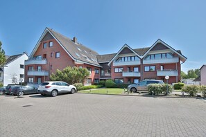 A multi-story residential building with a brick facade, multiple balconies, and a parking area with several cars.