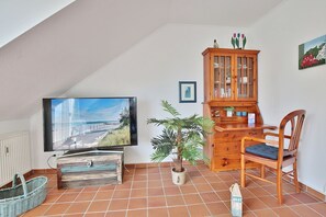 Una sala de estar con televisión, un mueble de madera, una silla, una planta en maceta y piso de baldosas.