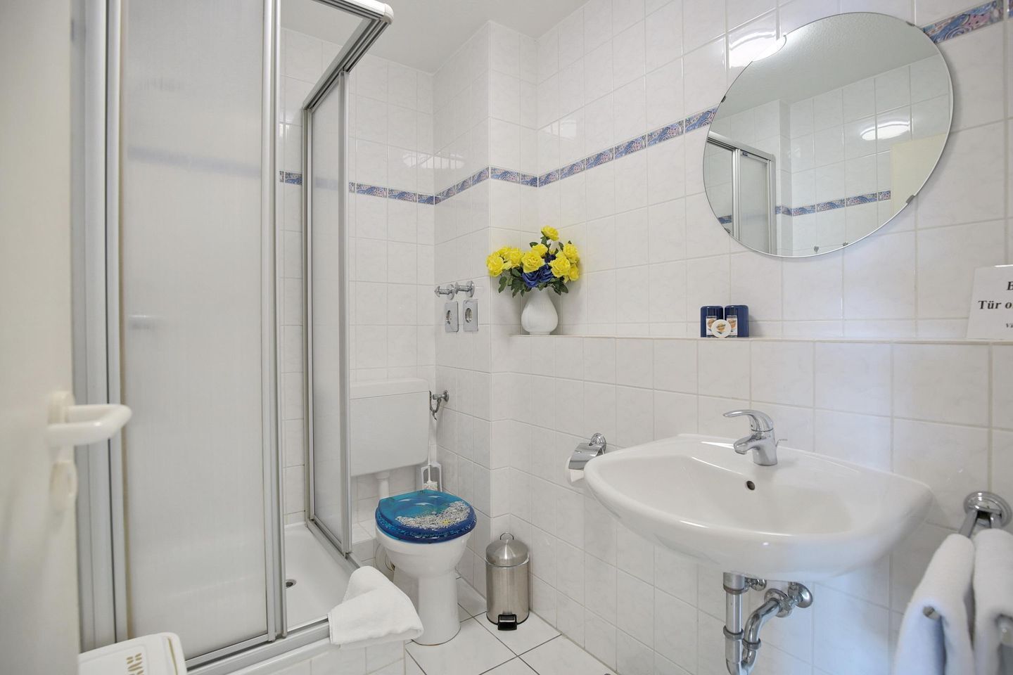 Ein Badezimmer mit Duschkabine aus Glas, einem weißen Waschbecken, einer Toilette mit blauem Deckel und einem Spiegel.