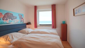 Une chambre d’hôtel avec un lit, une table de chevet, un tableau, une fenêtre avec des rideaux et une vue sur la mer.