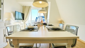 A dining room with a large table, chairs, a television, and a view of the city. - Strandschlösschen Appartement 15 - Strandschlößchen (Scharbeutz)