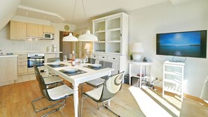A modern kitchen with a dining area, white cabinetry, and a flat-screen TV mounted on the wall. - Strandschlösschen Appartement 15 - Strandschlößchen (Scharbeutz)