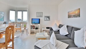 A modern living room with a grey sofa, wooden chairs, a TV, and artwork on the walls. - Strandschlösschen Appartement 5 - Strandschlößchen (Scharbeutz)