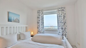 A bedroom with a bed, white bedding, a nightstand with a lamp, a painting on the wall, and a window with floral curtains. - Strandschlösschen Appartement 5 - Strandschlößchen (Scharbeutz)