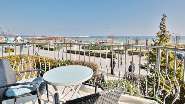 A balcony with a table and chairs, overlooking a coastal promenade with a view of the sea and a pier. - Strandschlösschen Appartement 5 - Strandschlößchen (Scharbeutz)