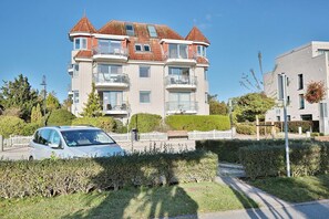Un immeuble résidentiel à plusieurs étages, doté de balcons, avec une voiture garée devant et un arbuste au premier plan.