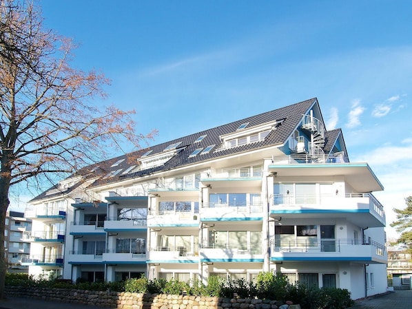 A multi-story residential building with balconies, a sloped roof, and a stone wall in front.