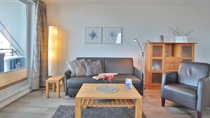 A modern living room with a grey sofa, a wooden coffee table, a leather armchair, and a wooden cabinet with glass doors. - Barque Apartment 404 - Barque (Scharbeutz)