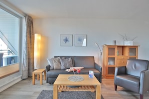 A modern living room with a grey sofa, a wooden coffee table, a leather armchair, and a wooden cabinet with glass doors. - Barque Apartment 404 - Barque (Scharbeutz)