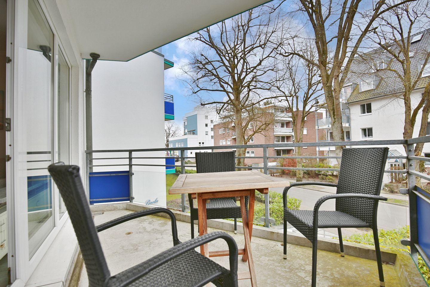 Ein Balkon mit zwei Korbstühlen und einem Holztisch, mit Blick auf eine Wohngegend mit Gebäuden und kahl stehenden Bäumen.
