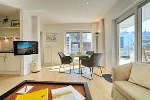 A modern living room with a sofa, a dining table with chairs, a TV mounted on the wall, and a window with a view of buildings. - Barke Apartment 324 - Barke (Scharbeutz)
