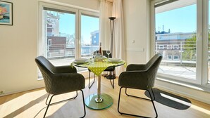 Un comedor con una mesa preparada para dos personas, dos sillas, una lámpara de pie y ventanas amplias con vistas a la ciudad.