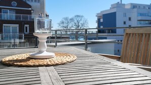 Un vaso de agua transparente sobre un pedestal blanco en una terraza de madera con un tapete tejido, con vistas a un cuerpo de agua y edificios.