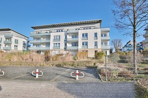 A multi-story residential building with balconies, a paved area in front, and a metal fence.