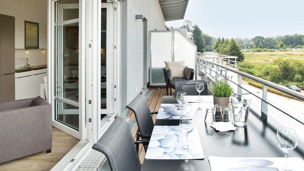 A modern outdoor dining area with a table set for two, a sofa, and a view of the countryside. - Strandglück Apartment 17C - Strandglück House C (Scharbeutz)