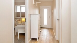 A narrow hallway with a white wardrobe, a bed with a checkered blanket, and a small table with a lamp. - Strandglück Apartment 17C - Strandglück House C (Scharbeutz)