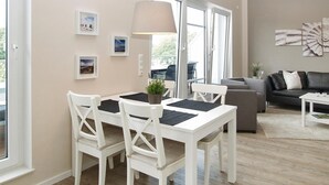 A modern living room with a white dining table, white chairs, a grey sofa, and a large window. - Strandglück Apartment 17C - Strandglück House C (Scharbeutz)