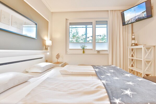 A hotel room with two beds, a TV mounted on the wall, a window with curtains, and a small table with a vase.