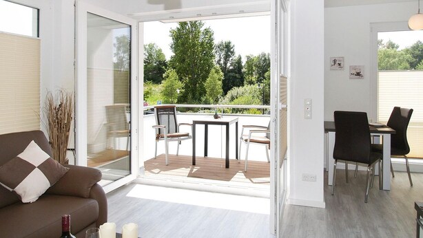 Uma sala de estar moderna com um sofá, uma área de jantar com cadeiras e uma mesa, e uma varanda com vista para o verde.