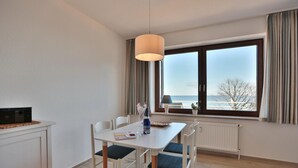 Ein moderner Essbereich mit einem weißen Tisch, blauen Sitzkissenstühlen, einem Radio und Meerblick durch große Fenster.
