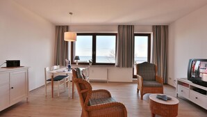 Ein modernes Wohnzimmer mit Essbereich, großen Fenstern mit Vorhängen, Rattanmöbeln und einem Fernseher.