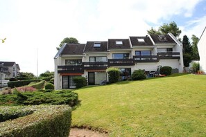 Um edifício residencial moderno de dois andares, com telhado plano, várias varandas e um gramado bem cuidado.