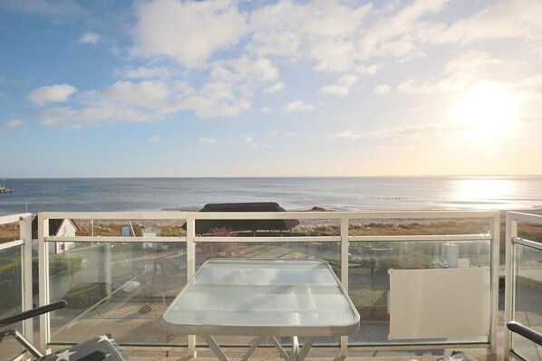 A balcony with a glass table and chairs overlooking a beach and ocean.