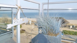 Blick aus einem Zimmer mit großem Fenster auf einen Strand, ein Schild mit der Aufschrift 'BEACH' und ein Blumentopf mit langen, dünnen Blättern.
