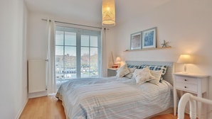 Ein Schlafzimmer mit Bett, Nachttischen, einer Pendelleuchte, einem Fenster mit Jalousien und Blick auf Gebäude.