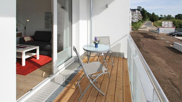 A balcony with a white wall, a sliding glass door, a small round table with a vase, and a red rug. - Strandglück, Apartment 8c - Strandglück House B (Scharbeutz)