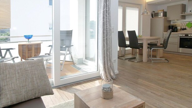 Una sala de estar moderna con un sofá, una mesa de centro y una zona de comedor con sillas y una mesa. Incluye una cocina con armarios y electrodomésticos.