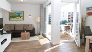 Una sala moderna con sofá, mesa de centro y televisor, que da a un balcón exterior con mesa y sillas.