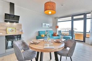 Una sala de estar moderna con una mesa de comedor redonda de madera, dos sillas grises tapizadas, un sofá azul y un ventanal con vistas a la ciudad.